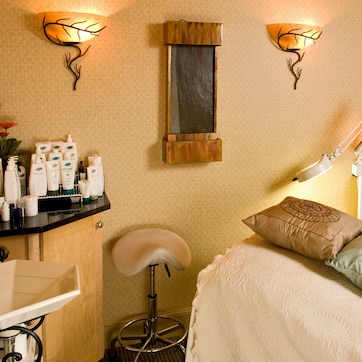 A well-lit room with a bed, a desk, and a chair. There are wall-mounted lights, a mirror, and a shelf with various items.