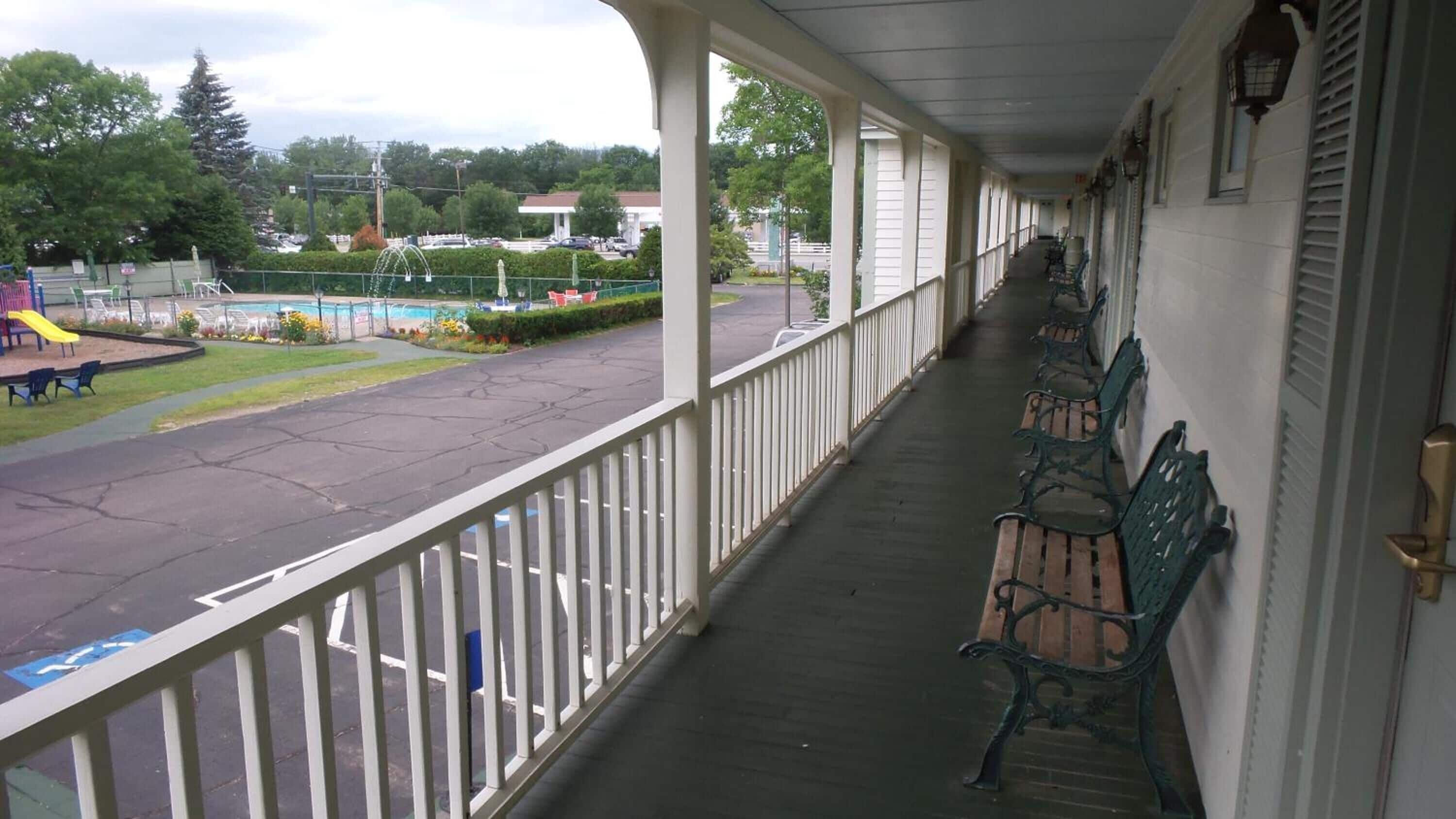 A covered porch with a row of wooden benches and a view of a swimming pool and playground.
