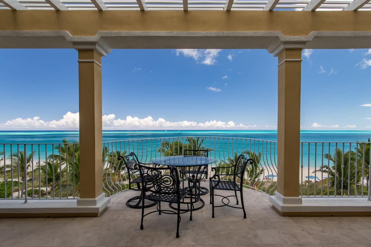 Ein Balkon mit Tisch und Stühlen, der auf ein türkisblaues Meer und Palmen blickt.
