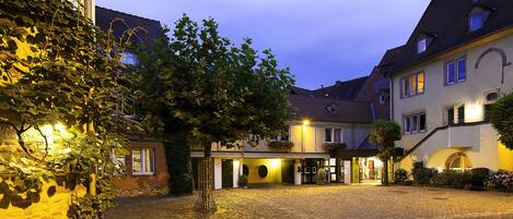 A cobblestone courtyard with buildings, a tree, and a well-lit entrance.