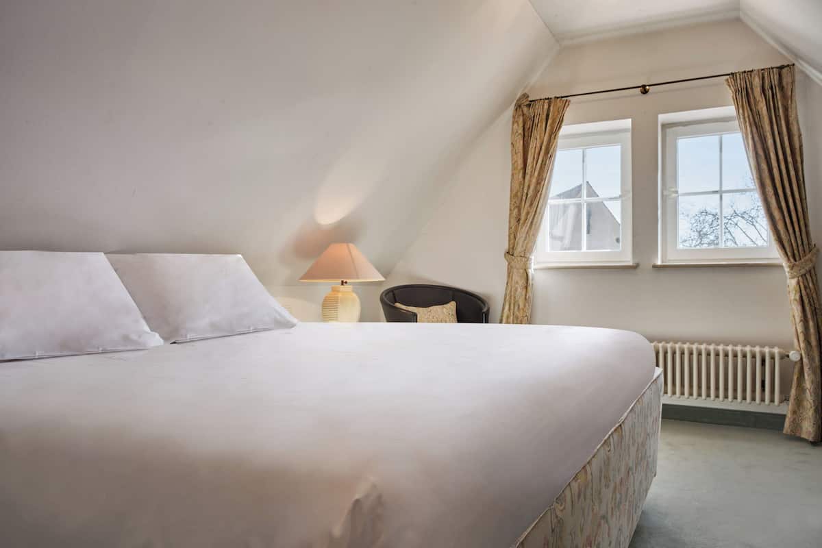 Un dormitorio con una cama grande, una lámpara de noche, una silla y una ventana con cortinas.
