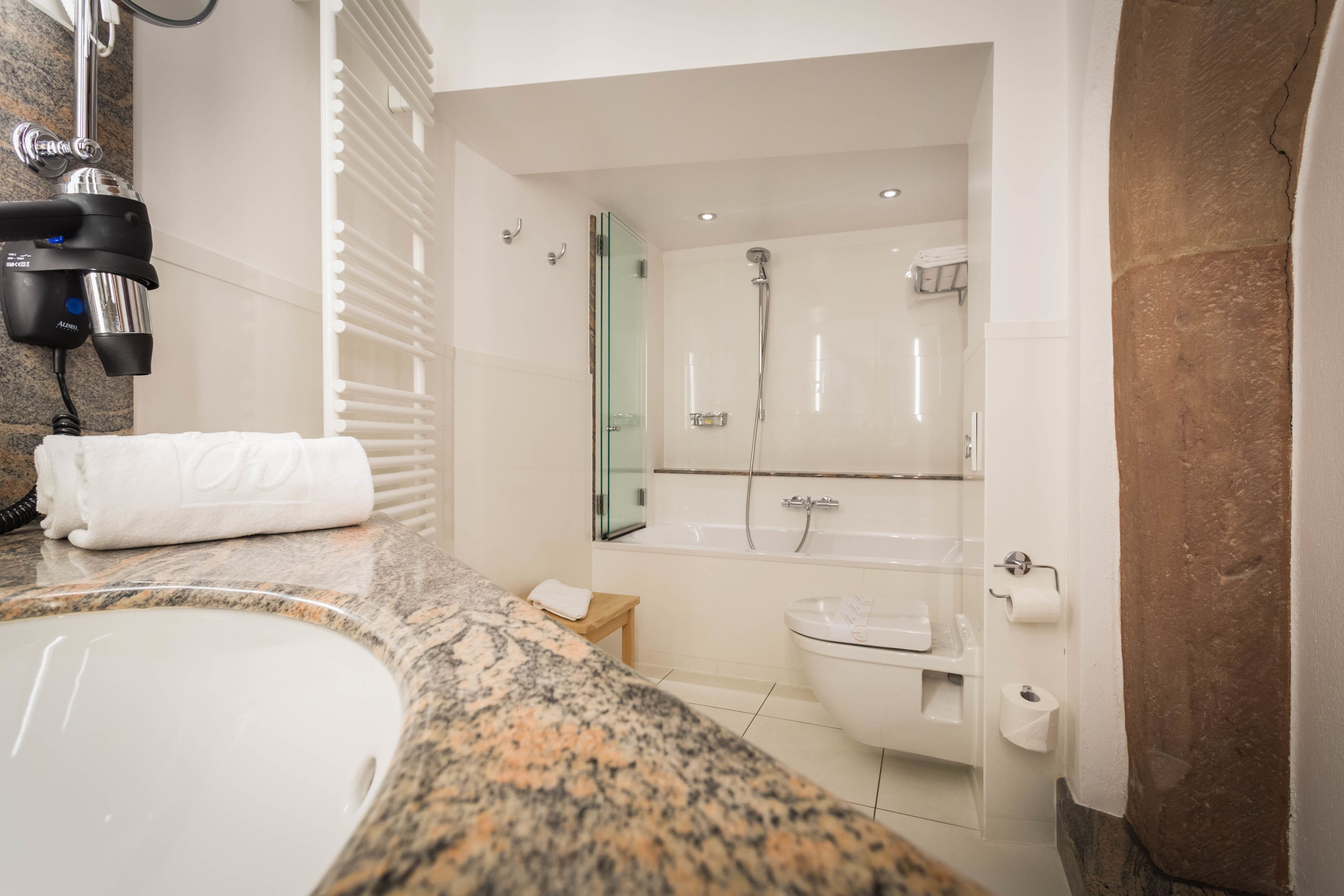 A modern bathroom with a granite countertop, a white sink, a glass-enclosed shower, and a toilet with a paper towel holder.