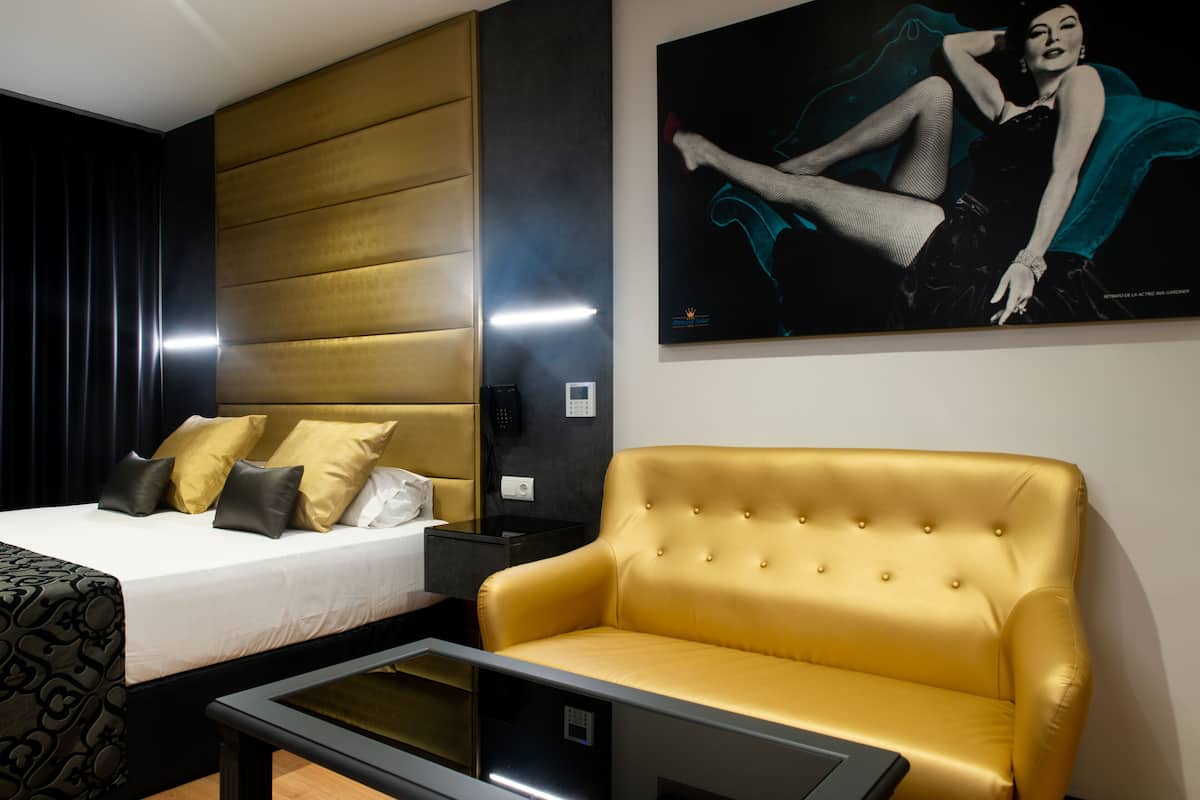 Hotellihuone, jossa on sänky, kullanvärinen sohva, yöpöytä ja juliste seinällä.