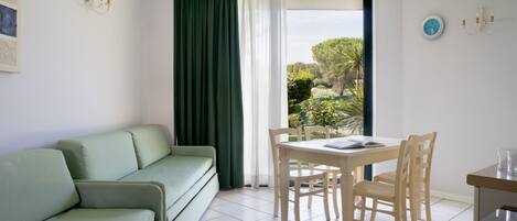 Een kamer met een groene bank, een eettafel met stoelen, een raam met gordijnen en een klok aan de muur.