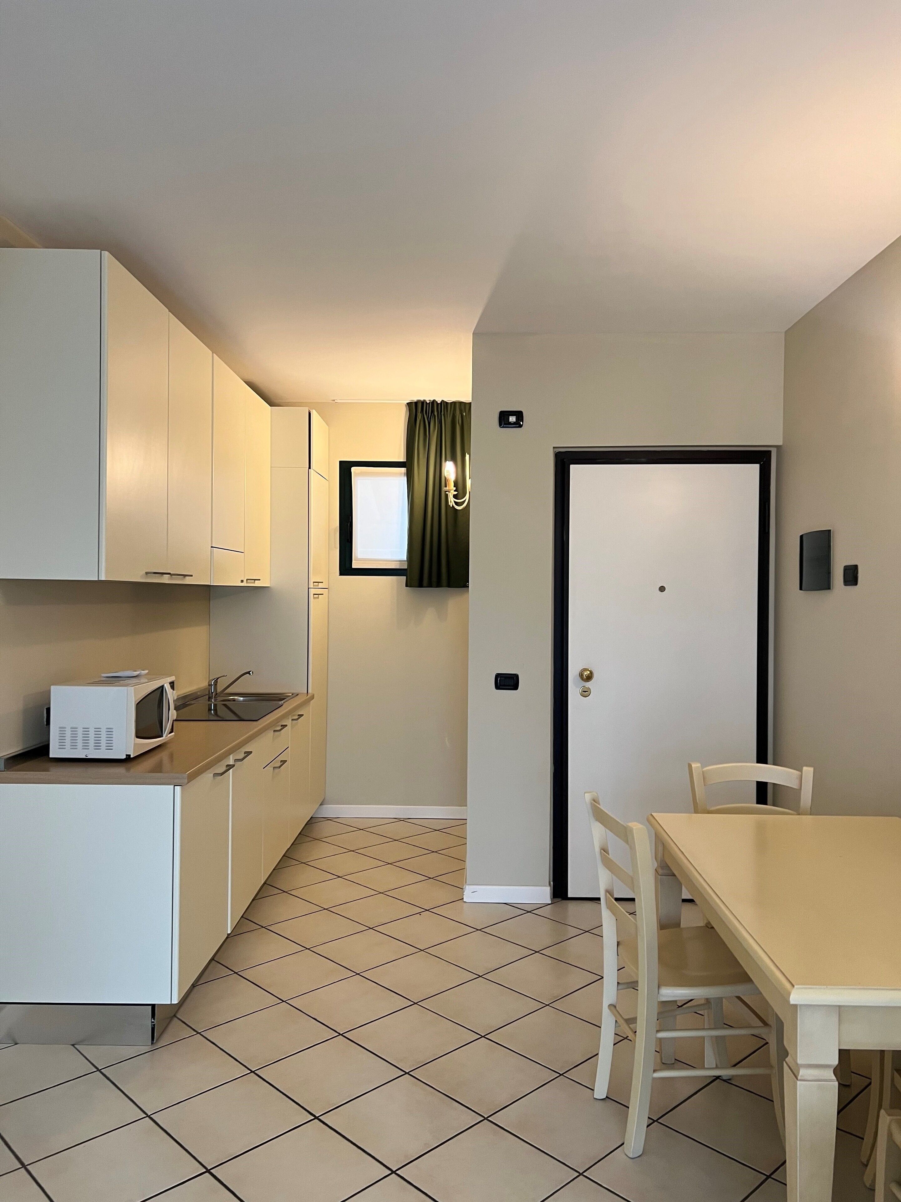 A compact kitchen with white cabinets, a microwave, and a sink. There is a dining table with chairs and a door with a handle.