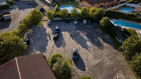 Vista aérea de un estacionamiento con varios coches, rodeado de árboles y edificios, incluyendo una piscina.