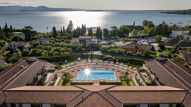 A resort with a central swimming pool, surrounded by sun loungers and umbrellas, with a view of a lake and mountains in the background.
