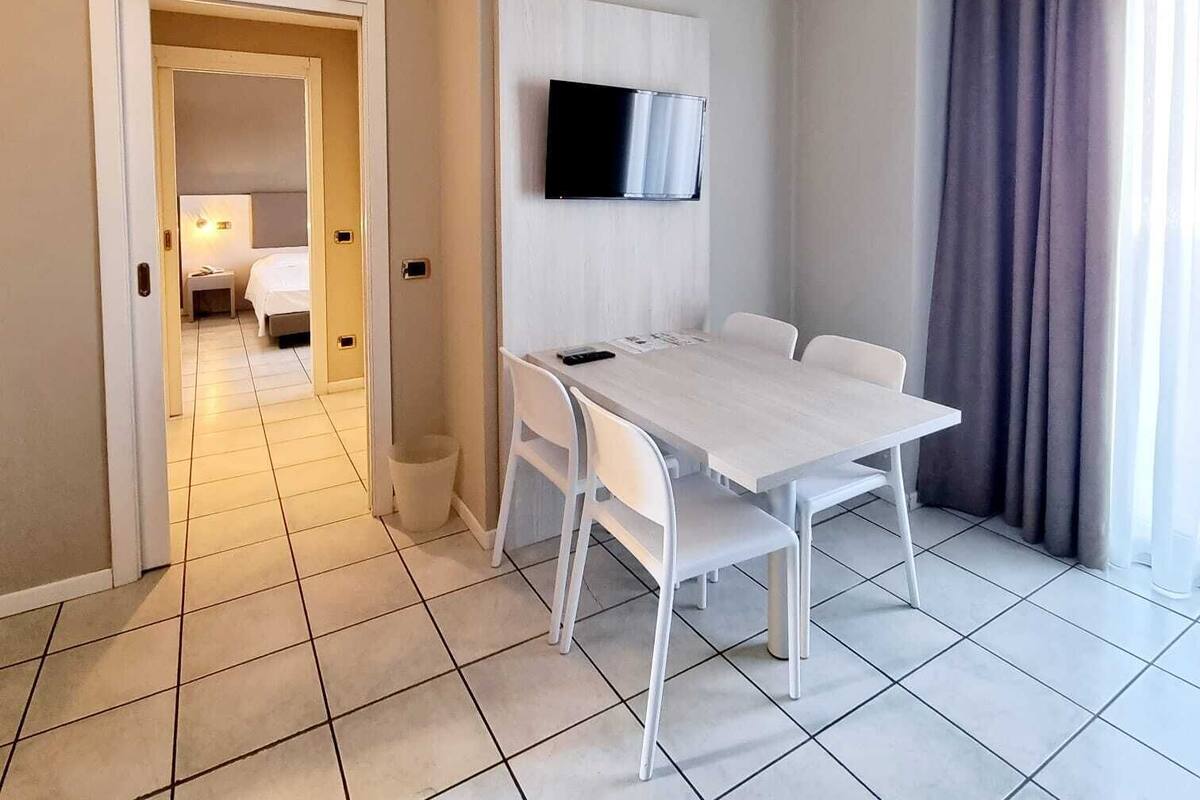 Ein Hotelzimmer mit einem weißen Tisch, Stühlen, einem an der Wand montierten Fernseher und Blick ins Freie durch ein Fenster mit Vorhängen.