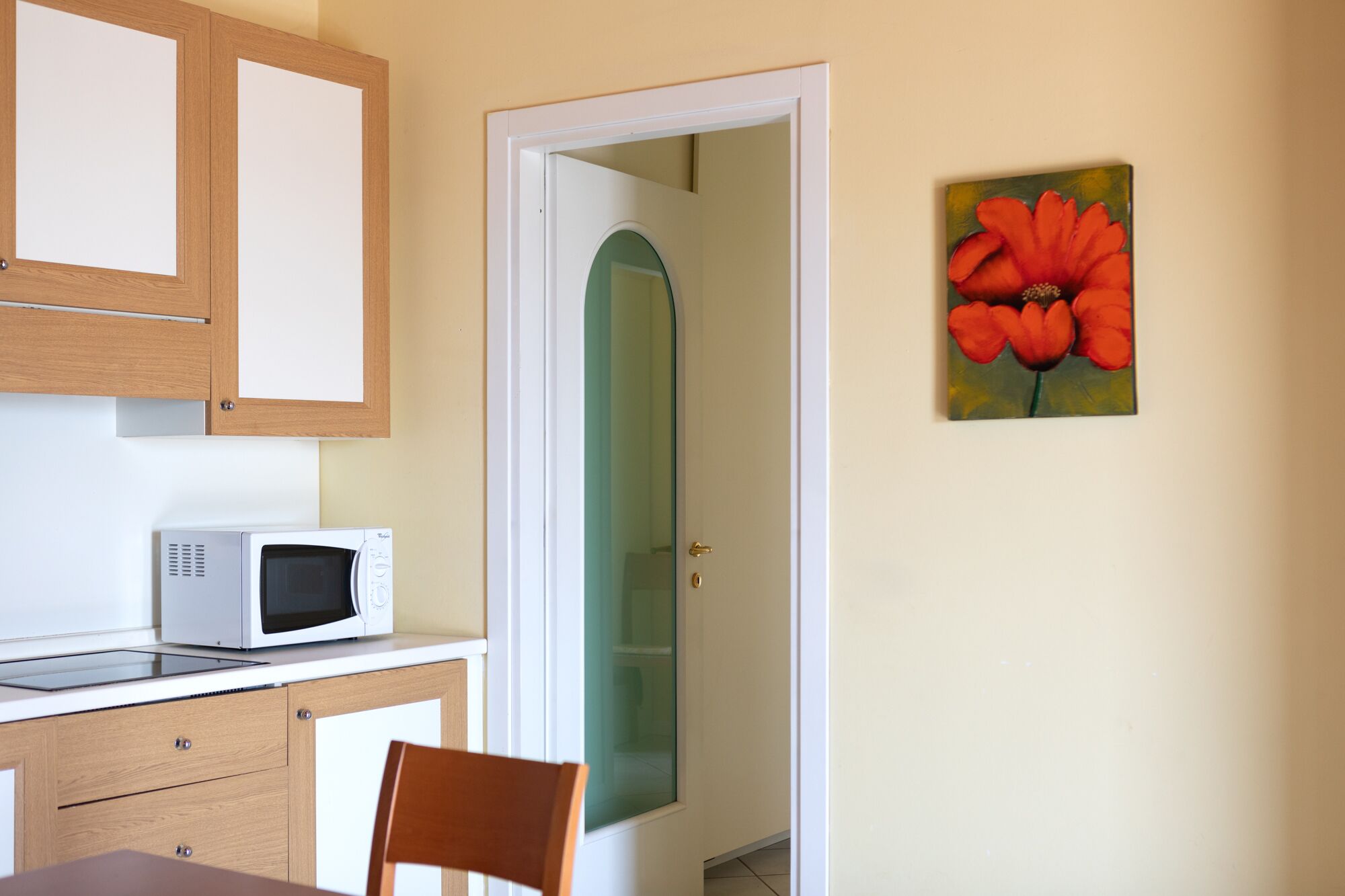 Una cocina con microondas, armarios de madera, una mesa de comedor con una silla y una puerta que da a otra habitación.