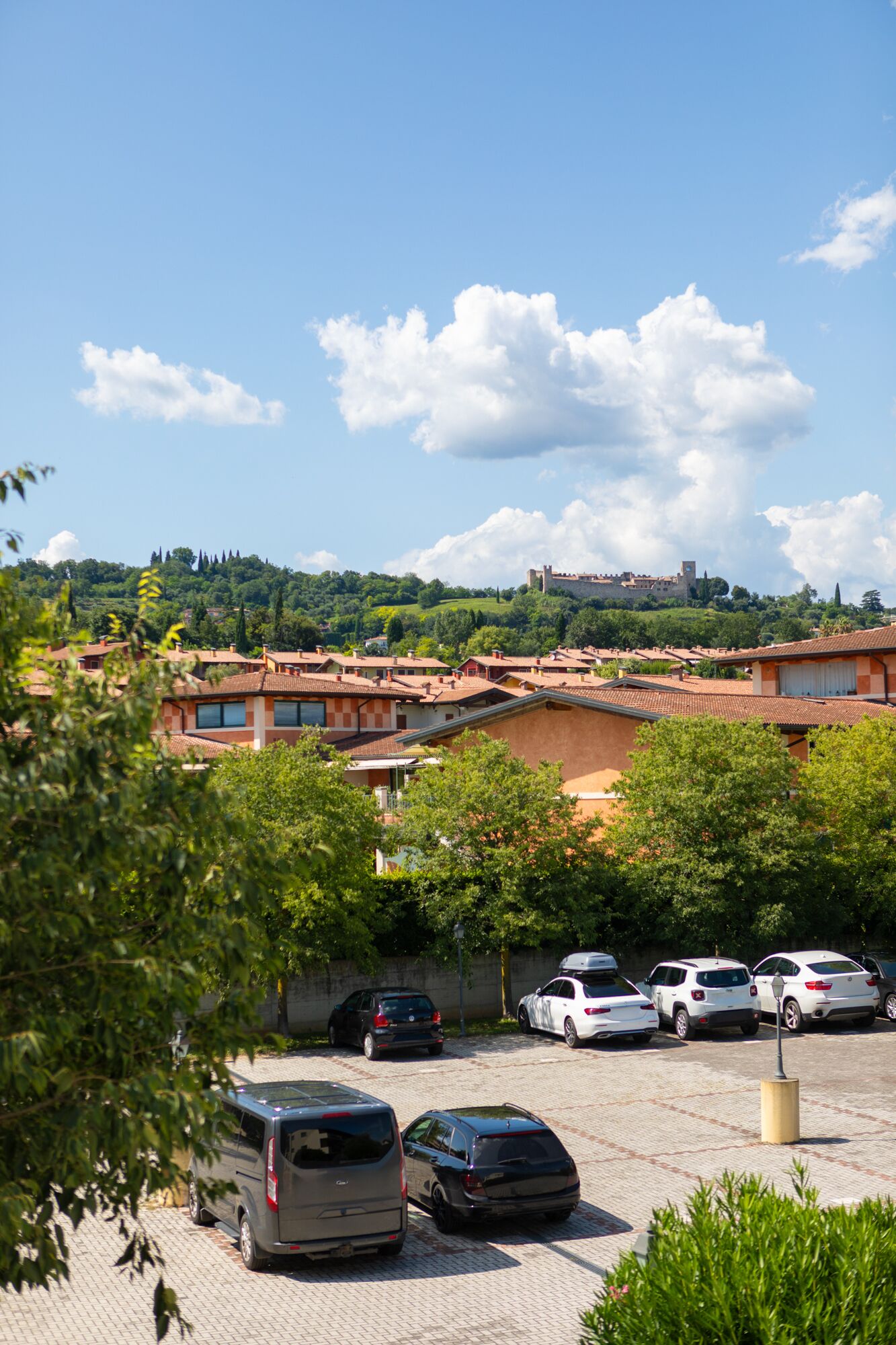 A parking lot with several cars, a building with a tiled roof, and a hill with trees in the background.