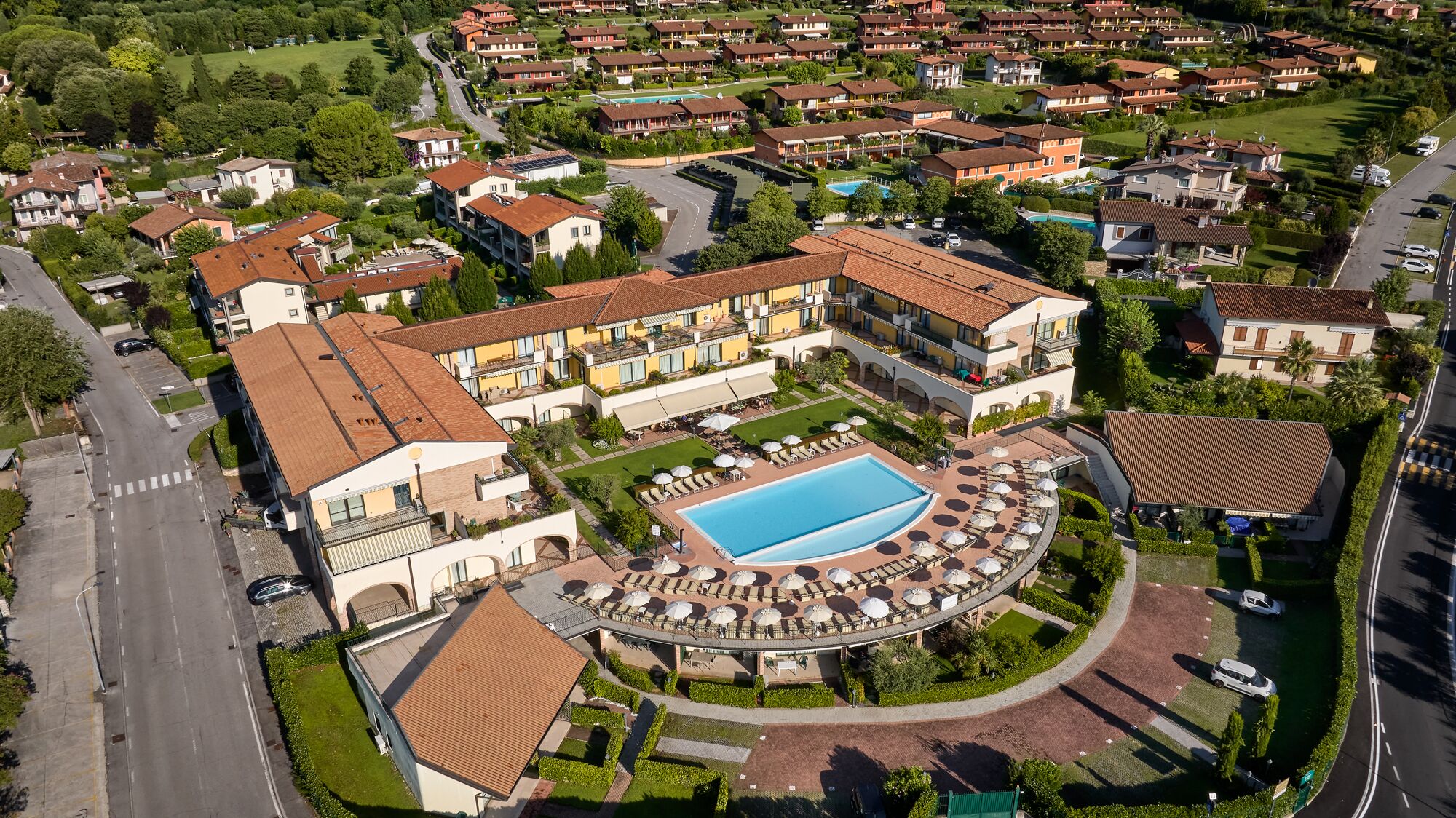 Une vue aérienne d’un quartier résidentiel avec une grande piscine entourée de chaises longues et de parasols, ce qui suggère qu’il s’agit d’un complexe hôtelier ou d’un hôtel.