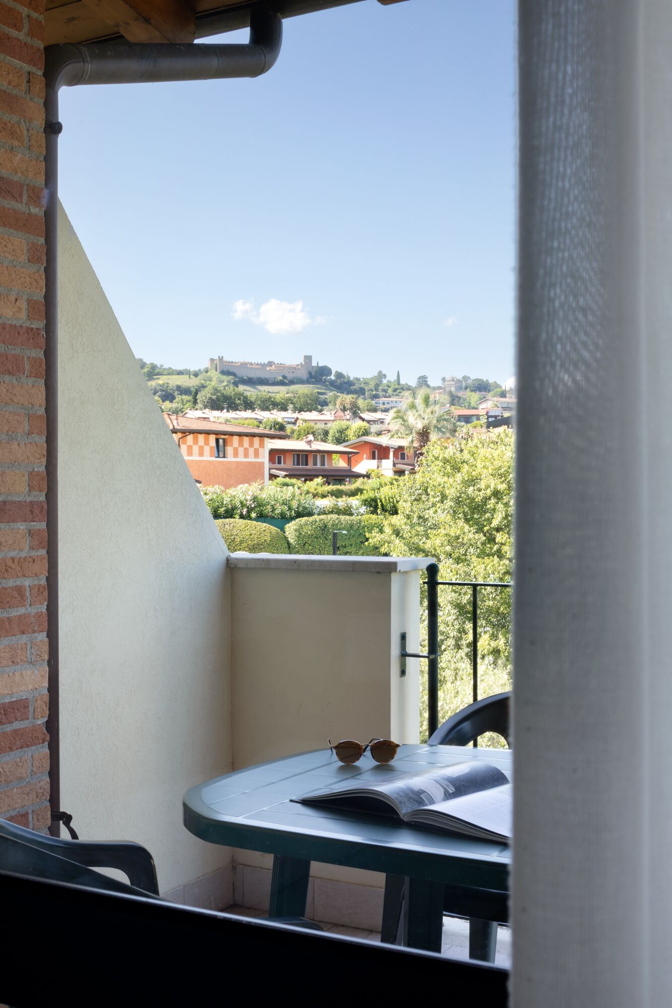 Un balcón con vistas a un pueblo, una mesa con un libro y gafas de sol, y un muro de ladrillo.