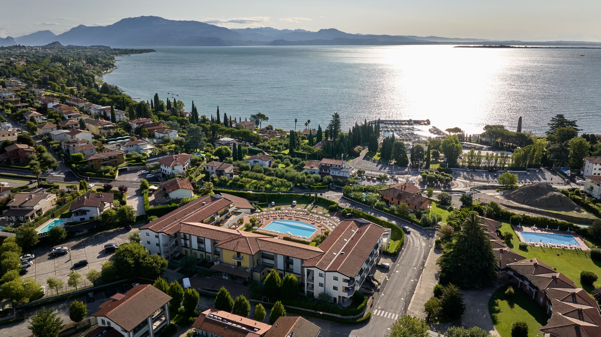 An aerial view of a coastal residential area with a large building featuring a swimming pool, surrounded by greenery and a marina.