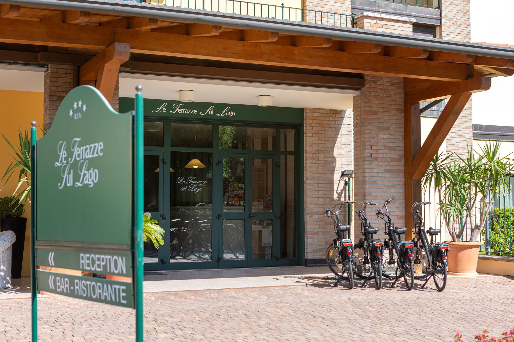 Ein Gebäudeeingang mit einem Schild, auf dem "Le Terrazze sul Lago" steht, und ein Empfangsbereich mit draußen abgestellten Fahrrädern.