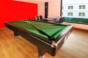 Una mesa de billar con bolas y tacos, una pared roja, una ventana con vistas a edificios y una mesa con sillas.