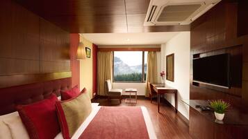 A hotel room with a large bed, a desk, a chair, a TV, and a window with a mountain view.