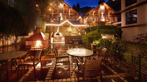 Un patio exterior acogedor con mobiliario de madera, un horno de pizza y una zona de comedor bien iluminada.