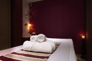 A neatly made bed with white linens and rolled towels, a striped bedspread, and a wall-mounted shelf with a vase and a candle.