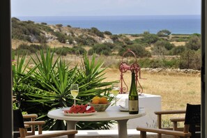 A table with a bottle of wine, glasses, a plate of watermelon, and a bowl of fruit, set against a backdrop of a coastal landscape. - Scala Apartments (Naxos)