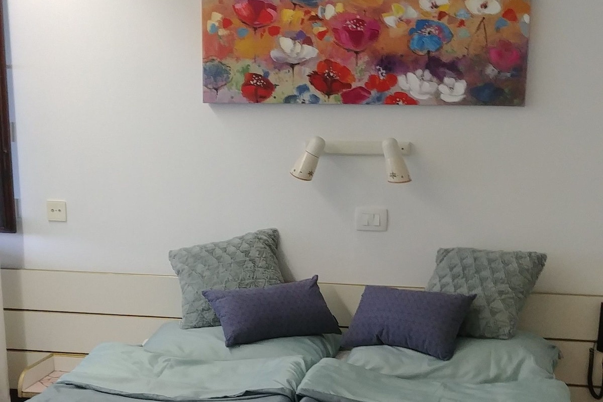Un dormitorio con cama, almohadas, una lámpara de pared y un cuadro floral.