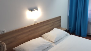 Habitación de hotel con cabecera de madera, dos camas blancas con almohadas, una lámpara de pared y una ventana con cortinas azules.