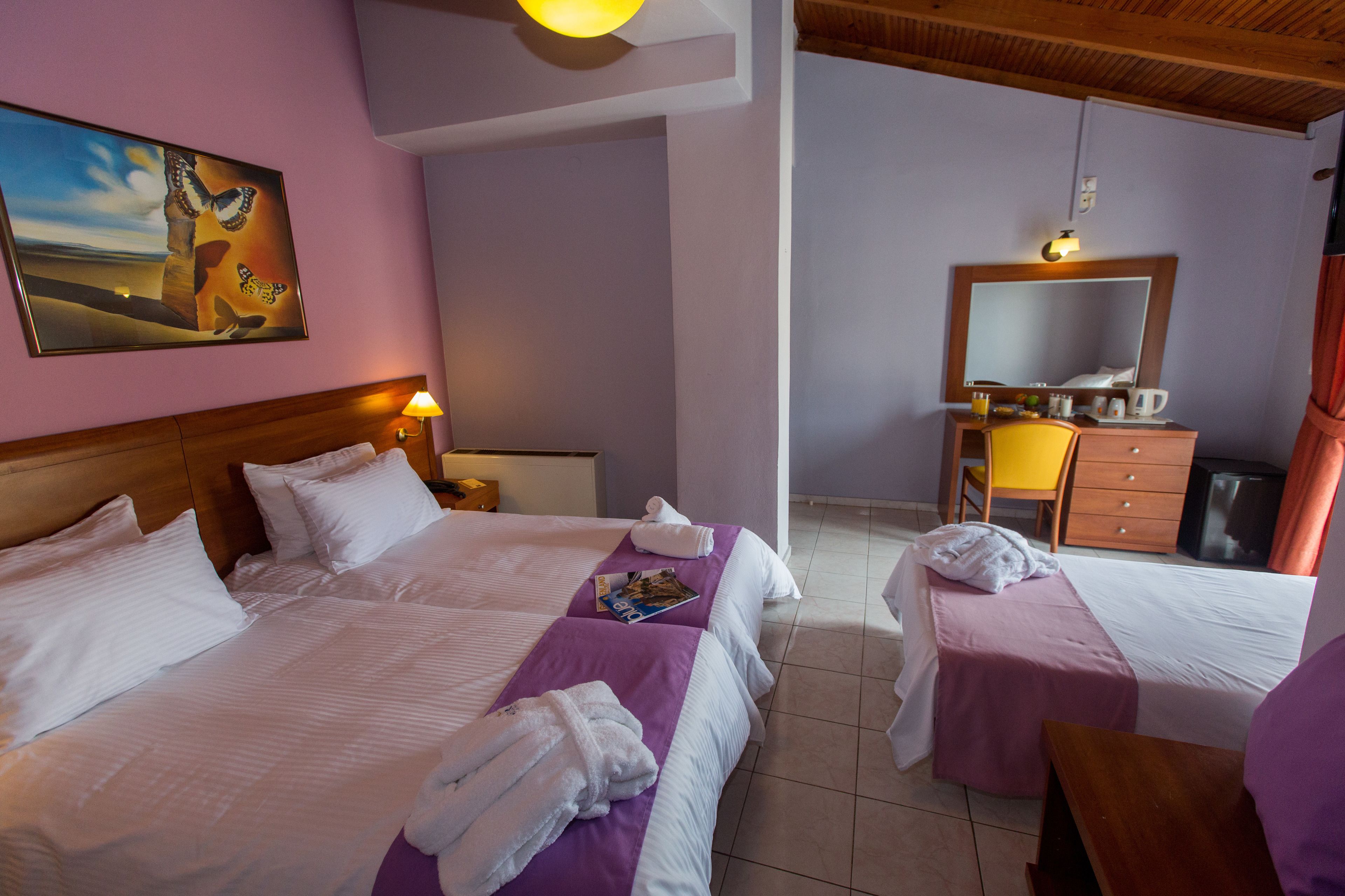 A hotel room with two beds, a wooden headboard, a painting, a desk with a chair, a mirror, and a small kitchenette.
