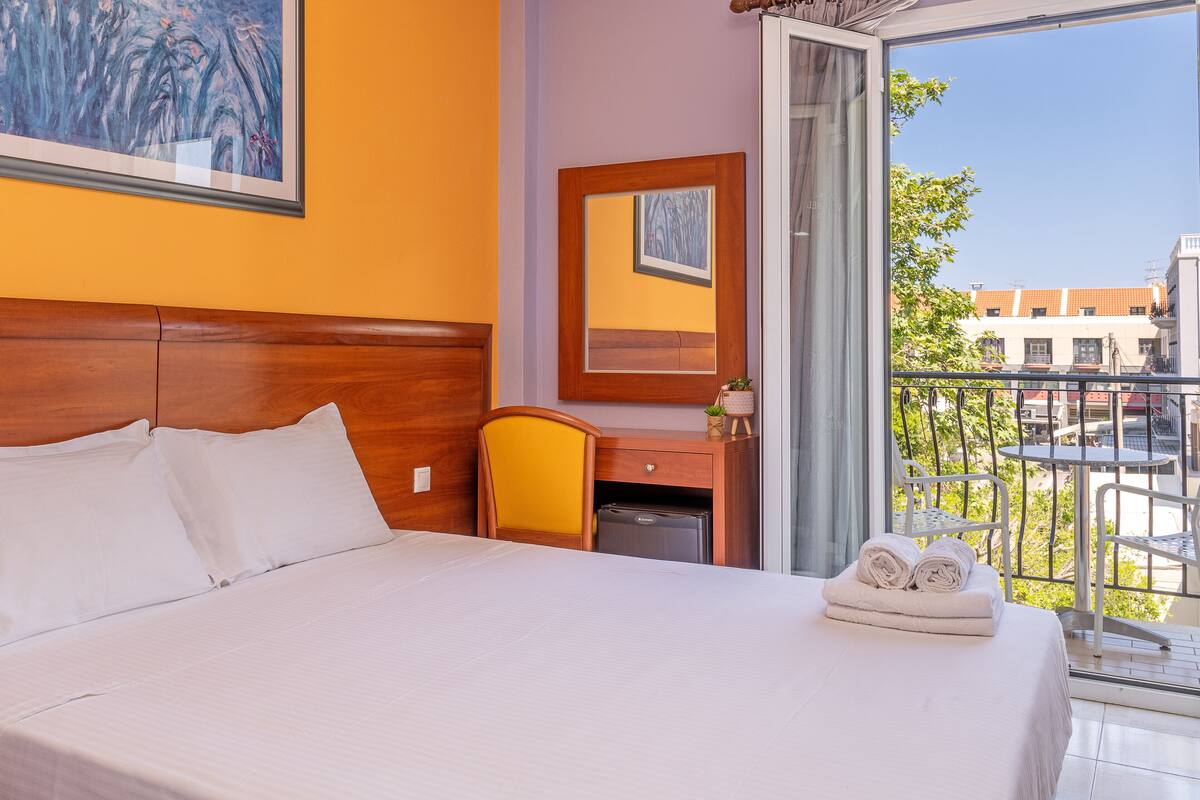 A hotel room with a bed, a desk, a chair, a small fridge, and a balcony with a view of buildings and trees.