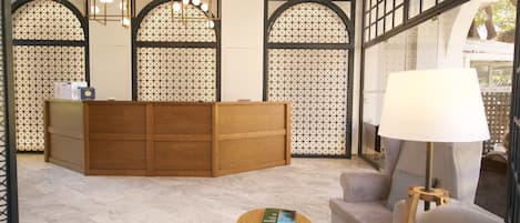A reception area with a wooden desk, a sofa, a small round table with a book, and a lamp.