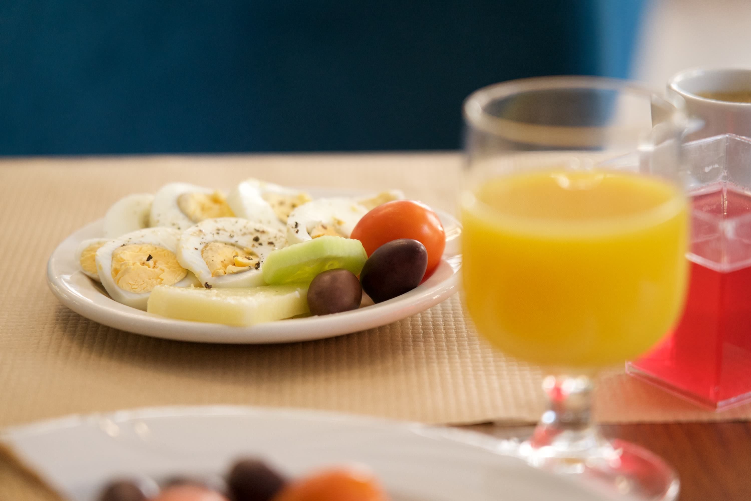 Mesa de café da manhã com prato de frutas fatiadas e ovos, um copo de suco de laranja e uma jarra de água.