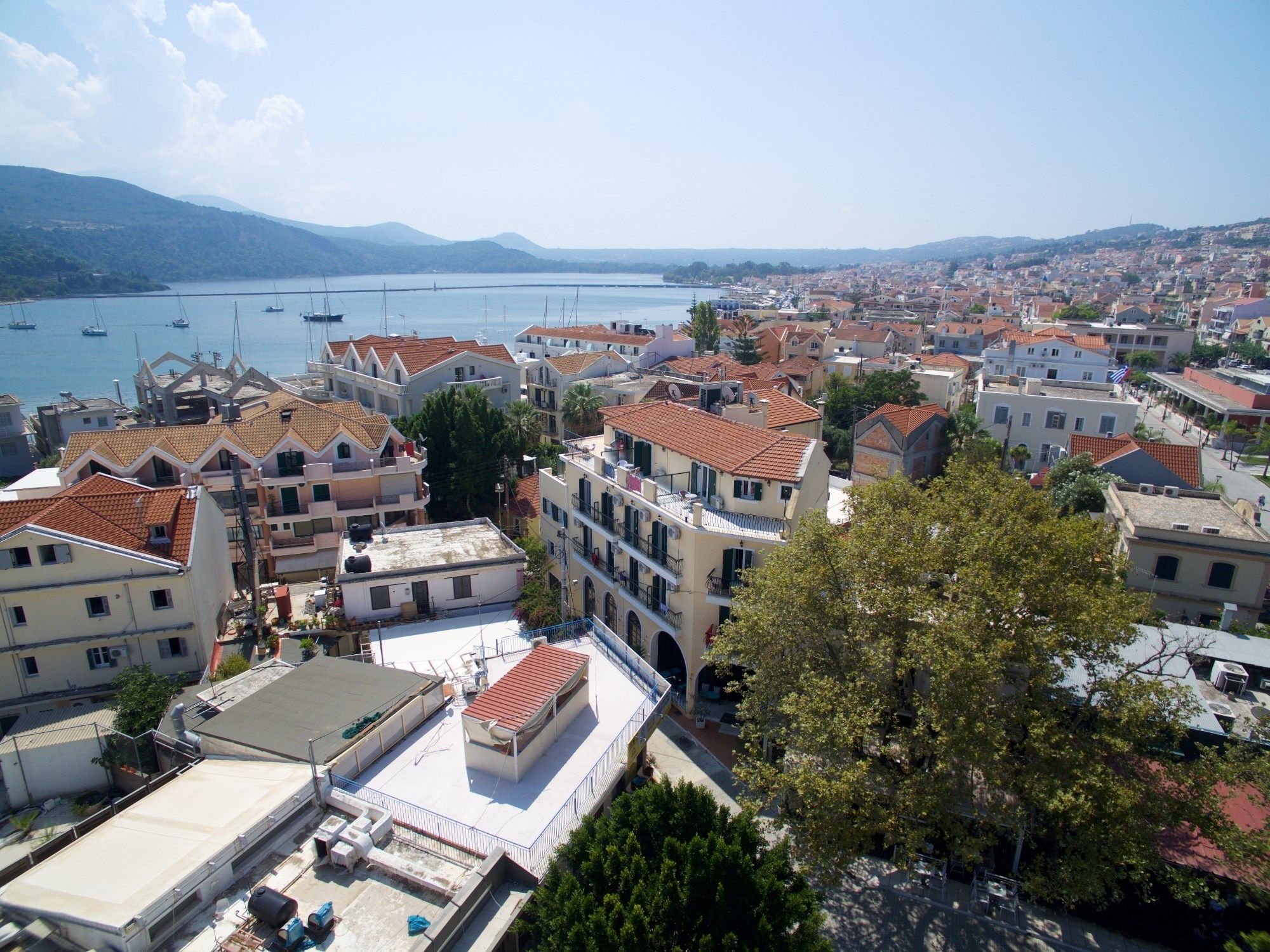 Un pueblo costero con edificios de tejas rojas, un puerto deportivo con barcos y una vista despejada del mar y montañas distantes.