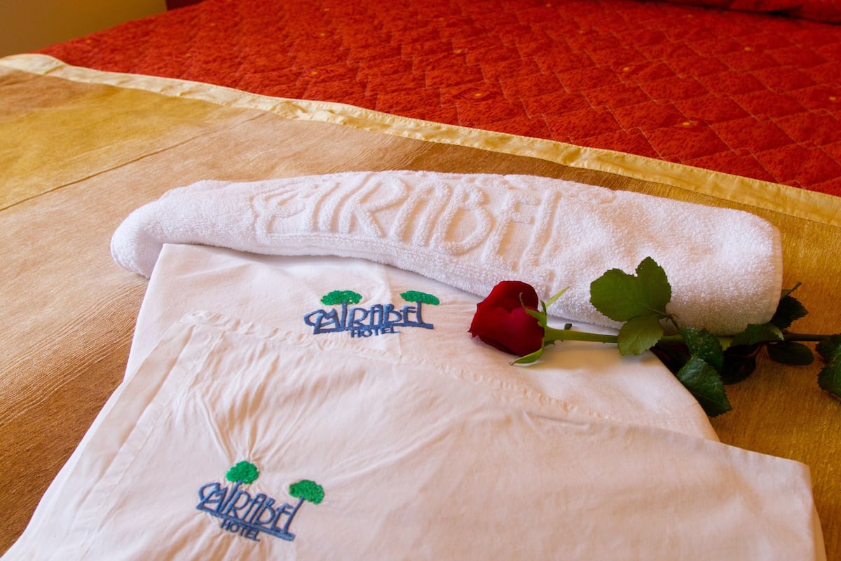 A neatly made bed with a red bedspread, a white towel with a logo, and a red rose on the bed.