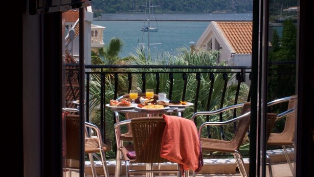 Un balcón con sillas de mimbre, una mesa con desayuno y vista a un velero en el agua.