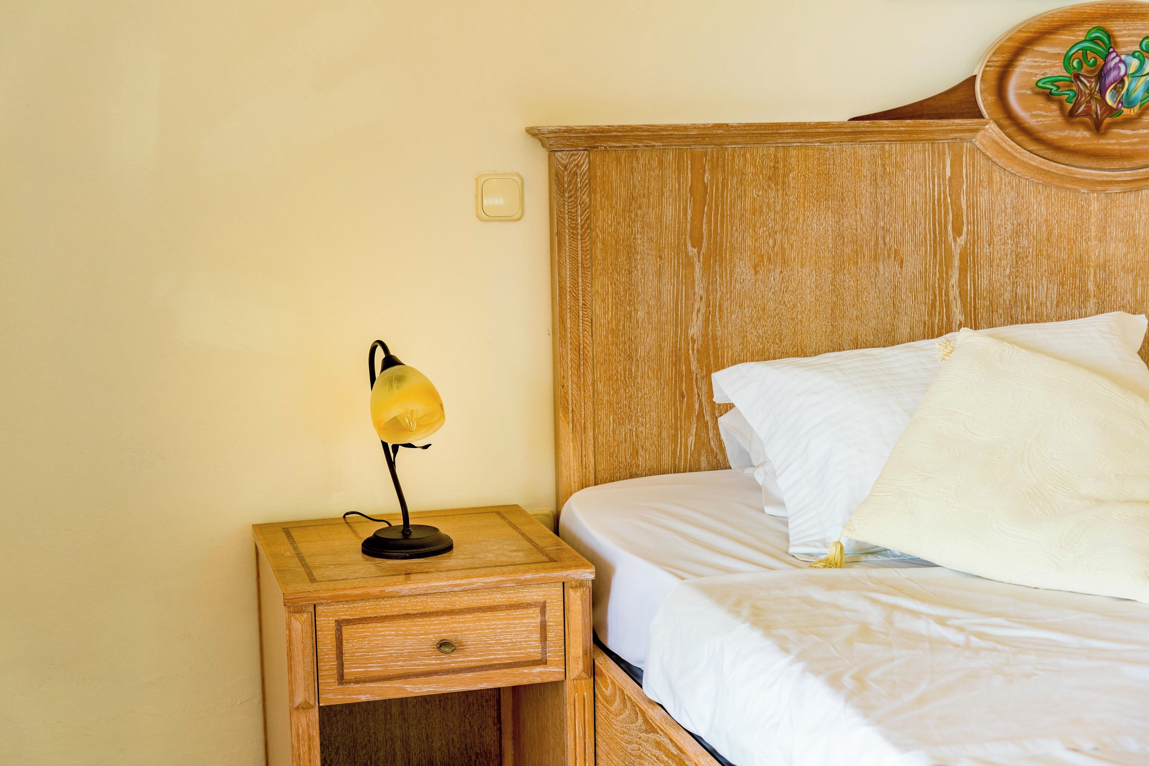 A neatly made bed with white linens, a wooden nightstand with a lamp, and a wooden headboard with a decorative crest.