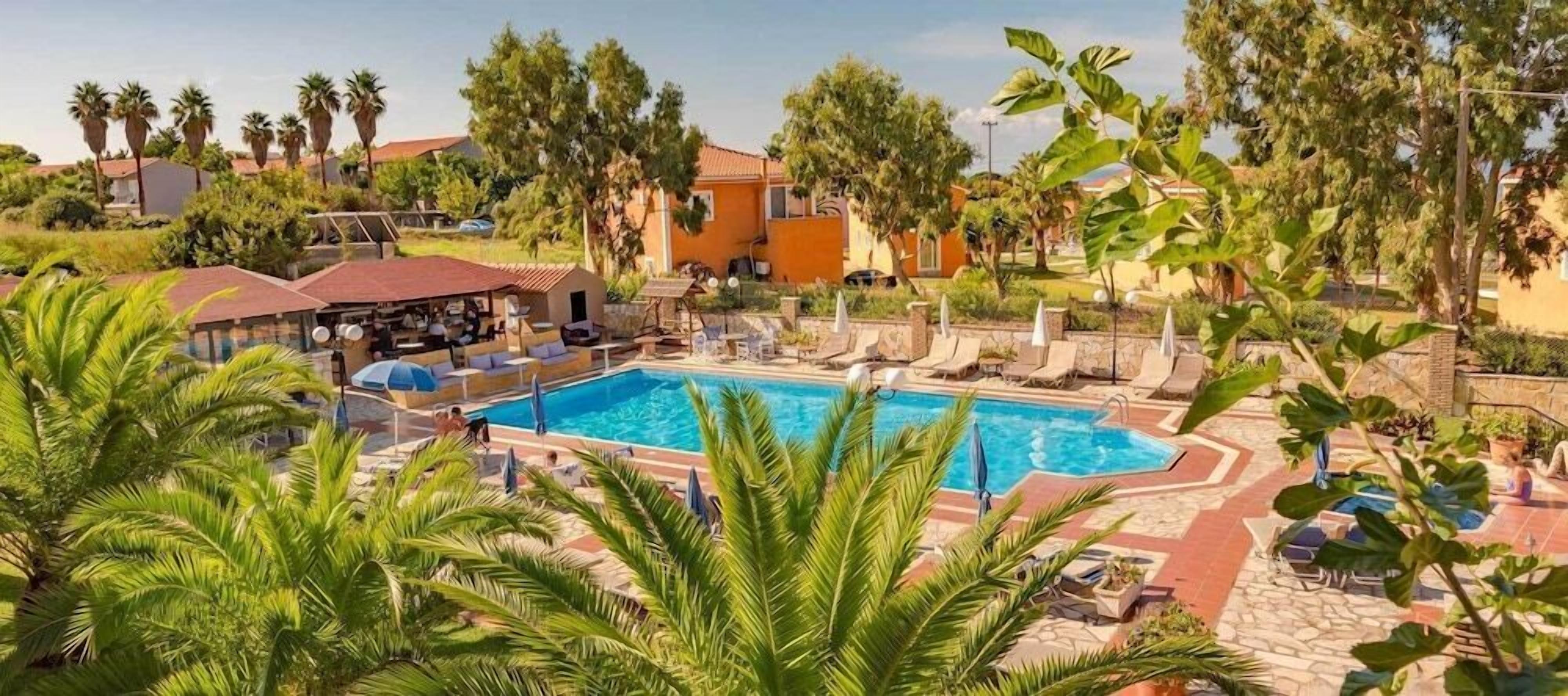 Zona de piscina con sillones de descanso y sombrillas, rodeada de palmeras y edificios.