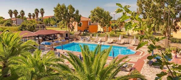 Zona de piscina con sillones de descanso y sombrillas, rodeada de palmeras y edificios.