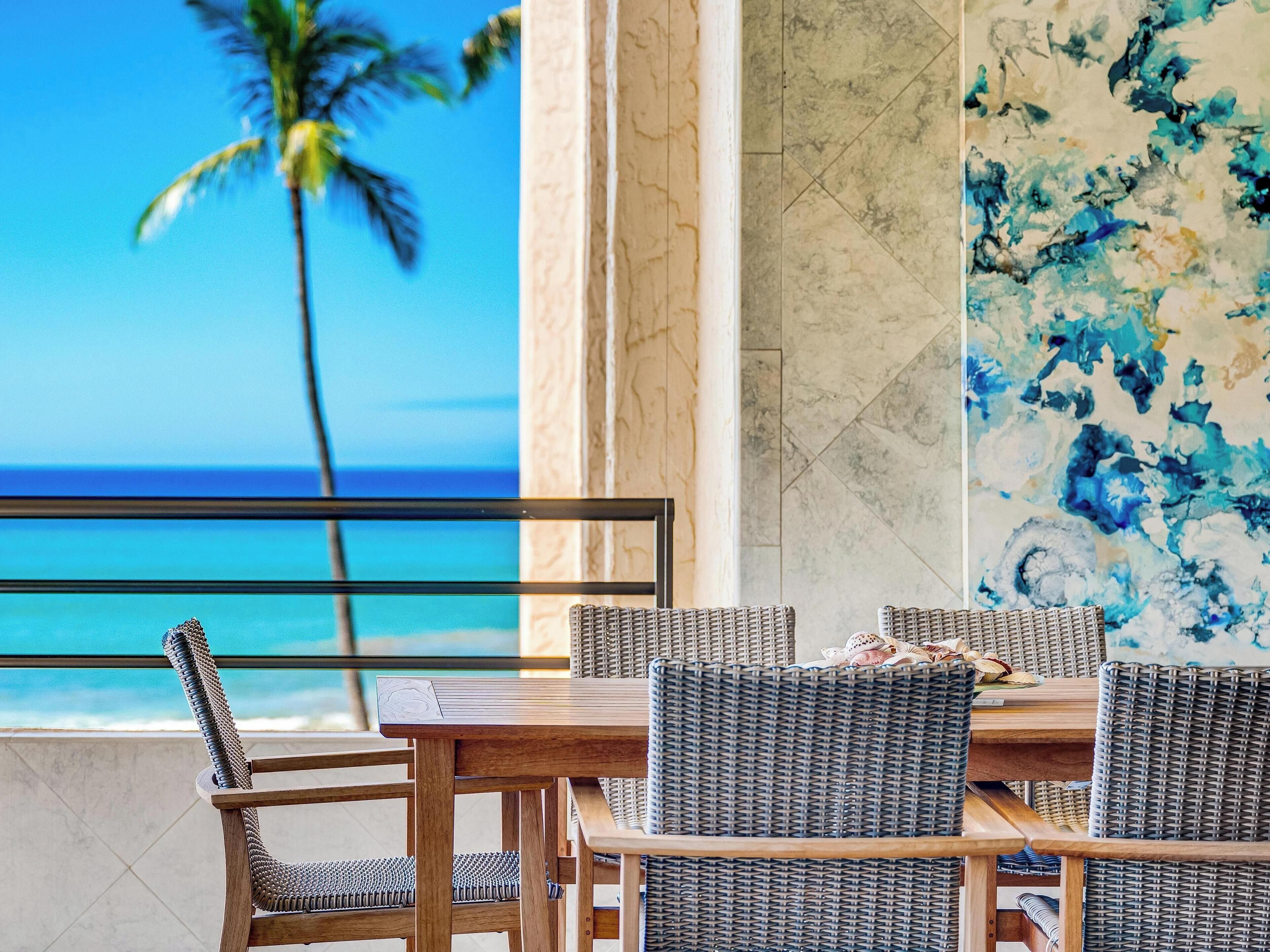 A dining area with wicker chairs and a wooden table, overlooking a beach with a palm tree.