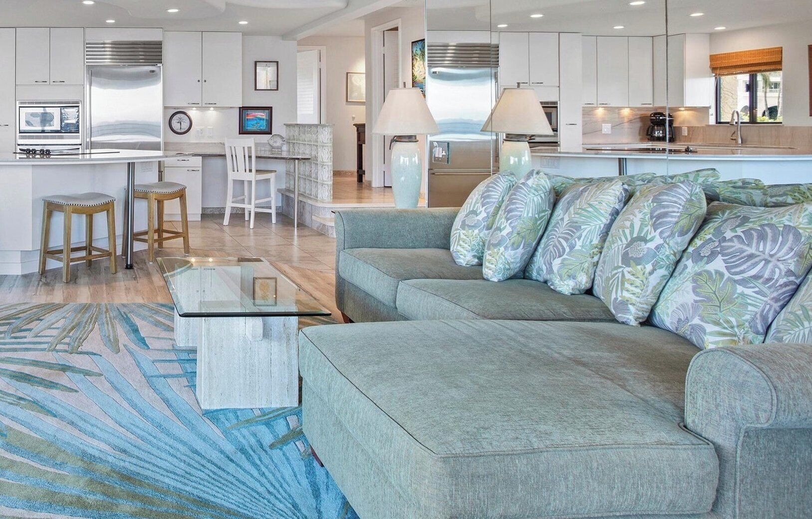 A modern kitchen with stainless steel appliances, a central island with stools, and a living area with a sofa and patterned cushions.