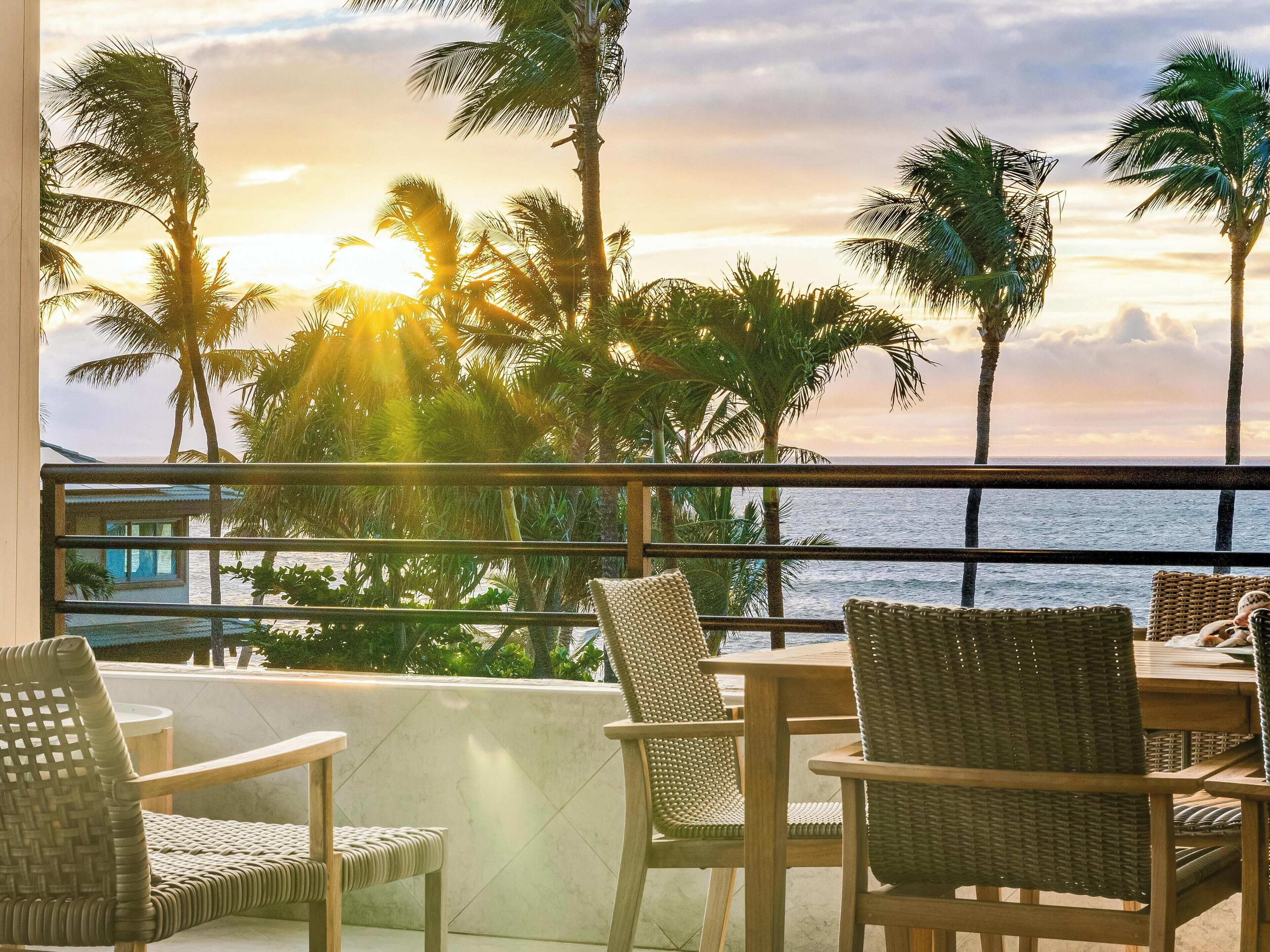 A balcony with wicker furniture, overlooking a palm tree-lined beach at sunset.