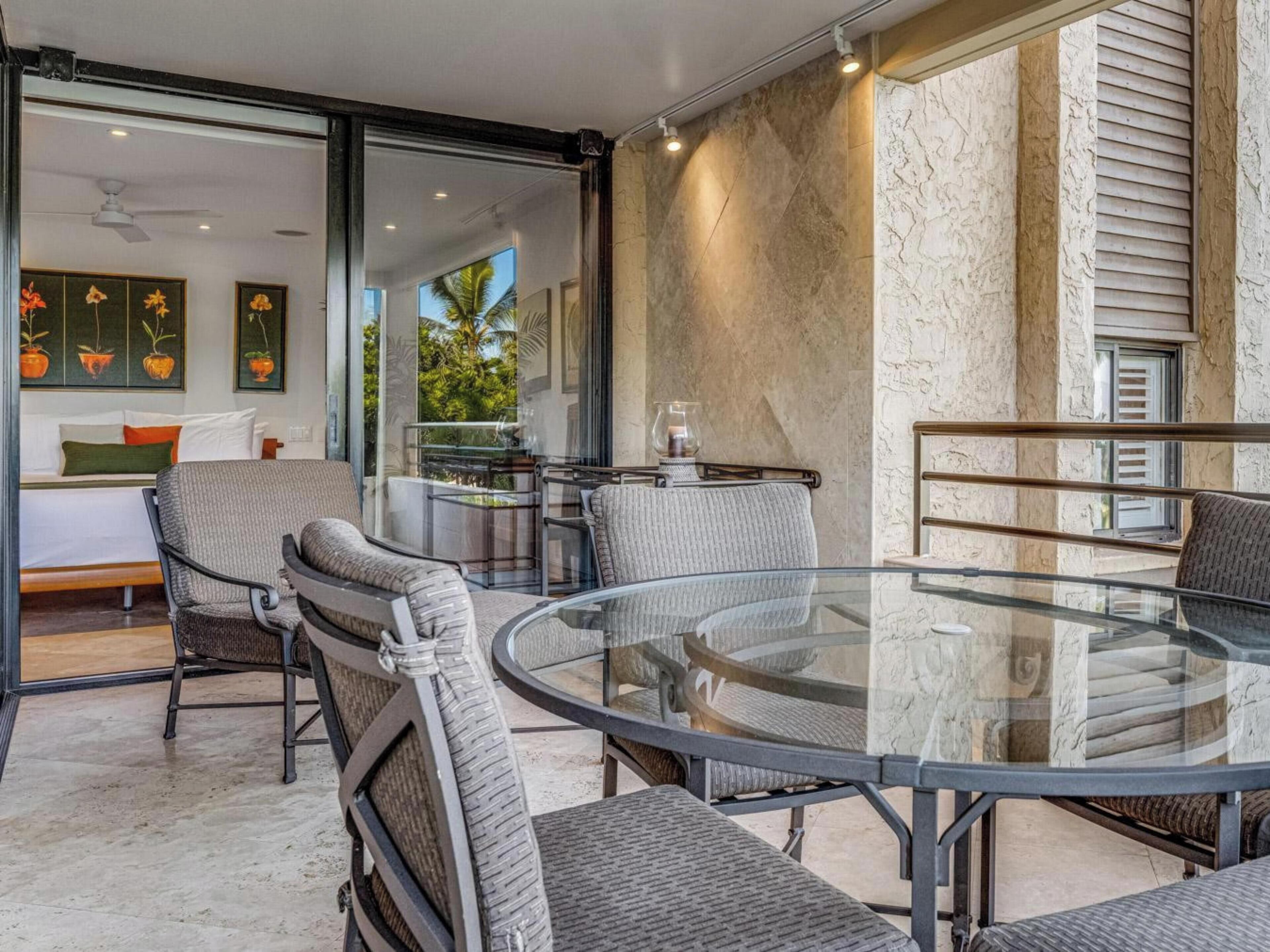 Un área de comedor moderna con una mesa de vidrio y sillas de metal, un sofá y un balcón con vistas a zonas verdes.