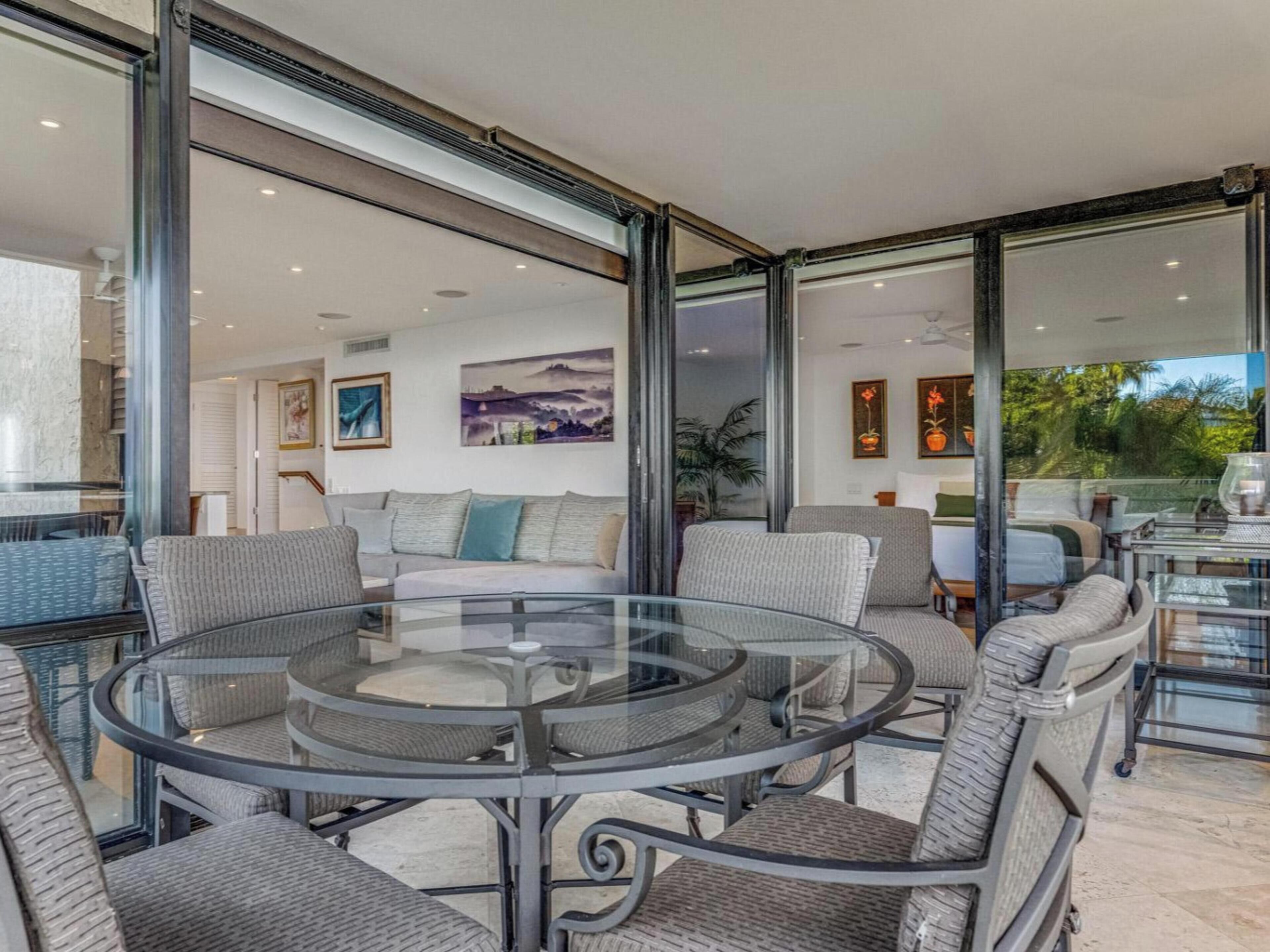 Un salon moderne avec une table en verre entourée de fauteuils en osier, un canapé et une grande fenêtre donnant sur l’extérieur.