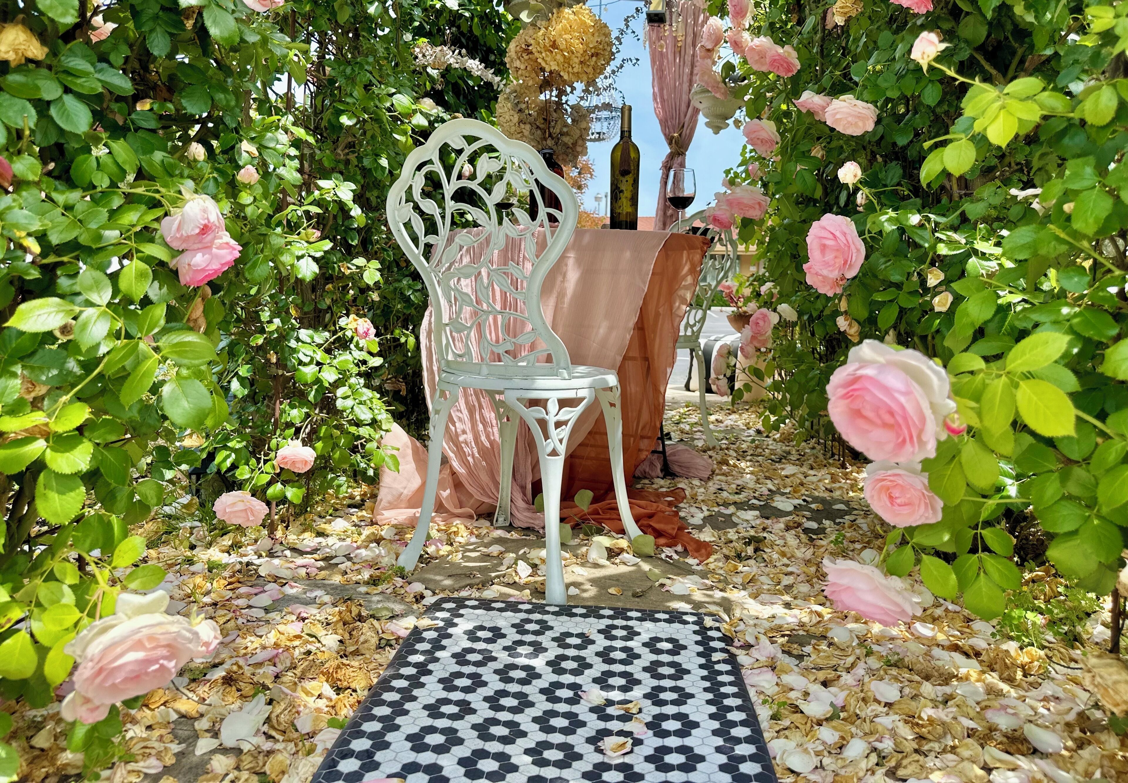Un jardín con una silla metálica blanca, una mesa con una botella y un vaso, rodeado de rosales y un suelo de baldosas.