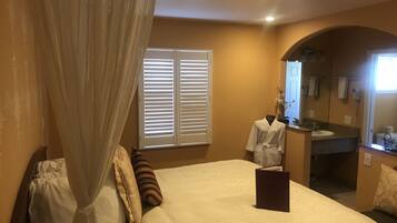 Un dormitorio con cama con dosel, una mesita de noche, un tocador, una ventana con persianas, un baño con lavamanos y espejo, y un arco de entrada.
