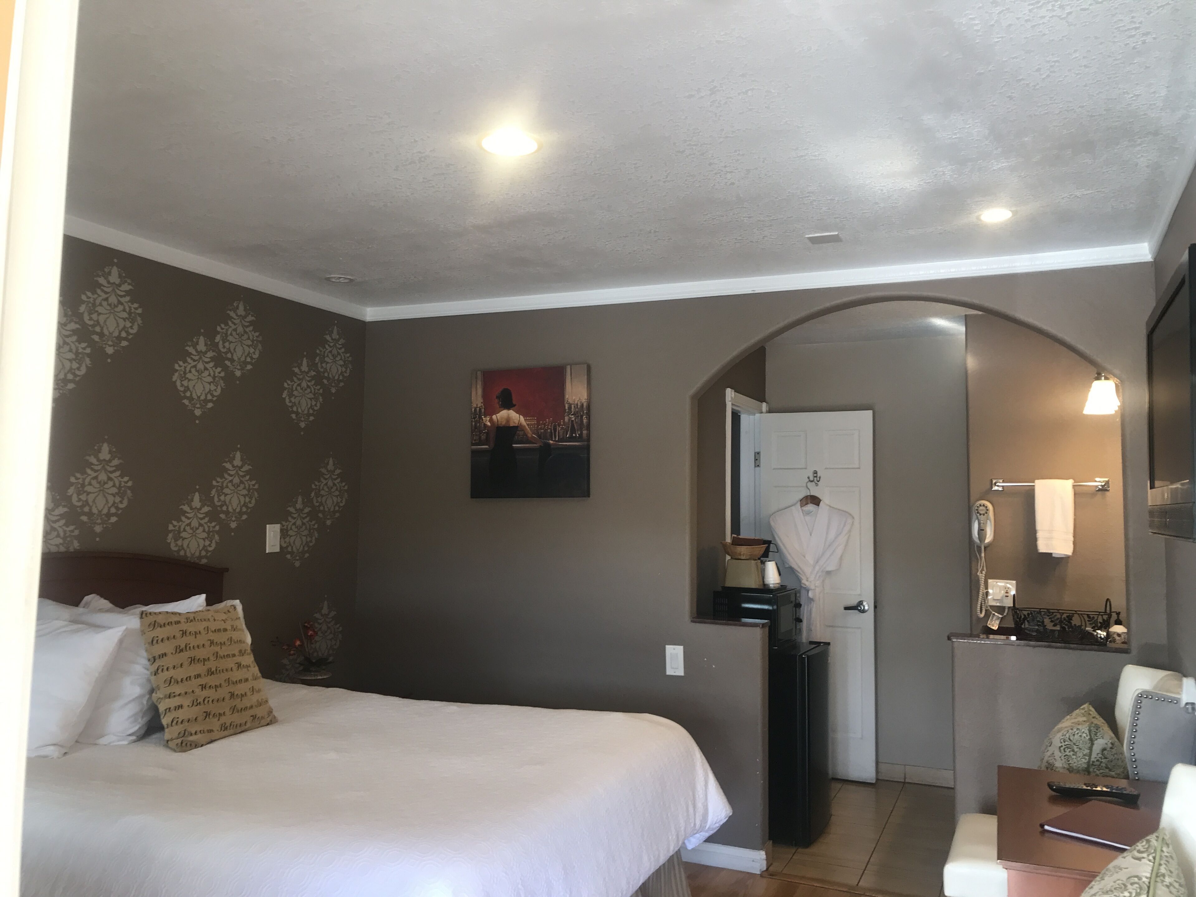 A hotel room with a bed, a desk, a chair, a mirror, a door, and a bathroom visible through an archway.