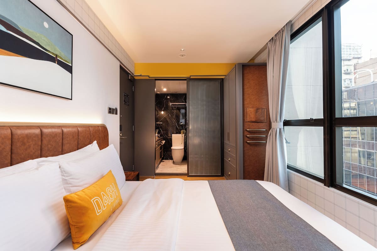 A hotel room with a bed, a large window, a bathroom, and a view of the city.