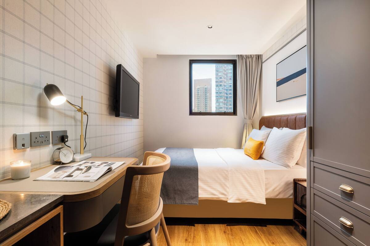 A hotel room with a bed, desk, chair, TV, and a window with a city view.