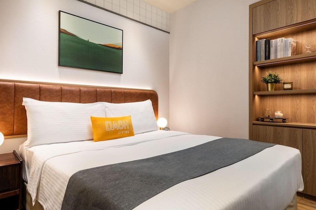 Una cama bien hecha con cabecera, una mesita de noche con lámpara, una estantería y un cuadro enmarcado en la pared.