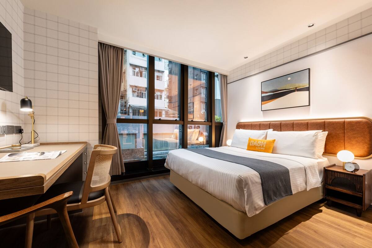 A hotel room with a large bed, a desk, a chair, a nightstand, and a window with a view of buildings.