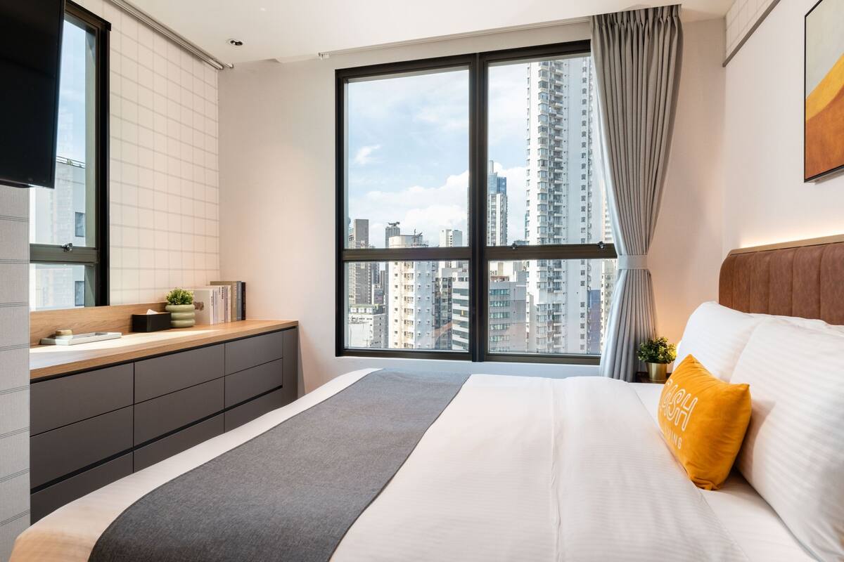 A modern hotel room with a large bed, a bedside table, a built-in bench, a window with a city view, and a flat-screen TV mounted on the wall.