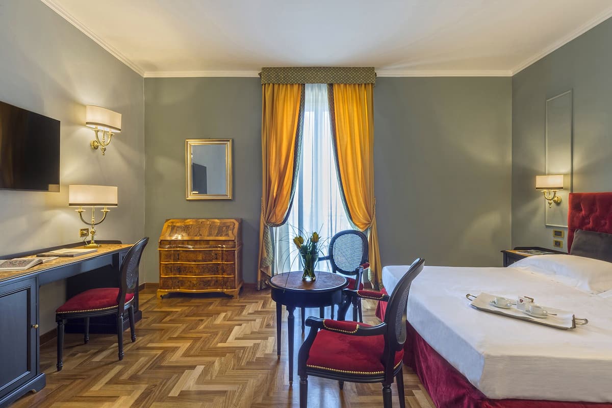 A hotel room with a bed, a desk with a chair, a small table with a chair, a dresser, and a television on the wall.