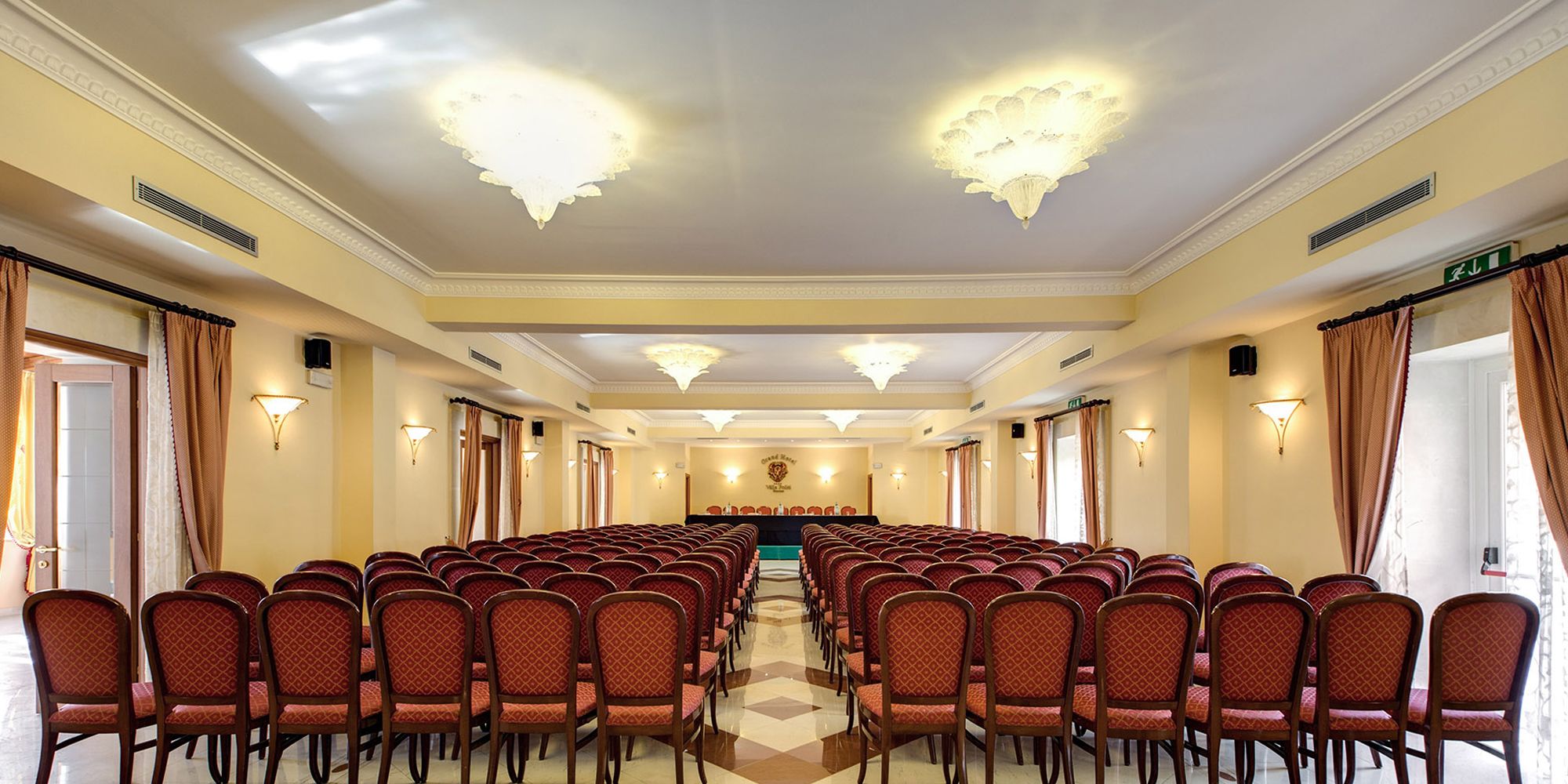 A conference room with rows of chairs facing a stage, a chandelier, and wall sconces.